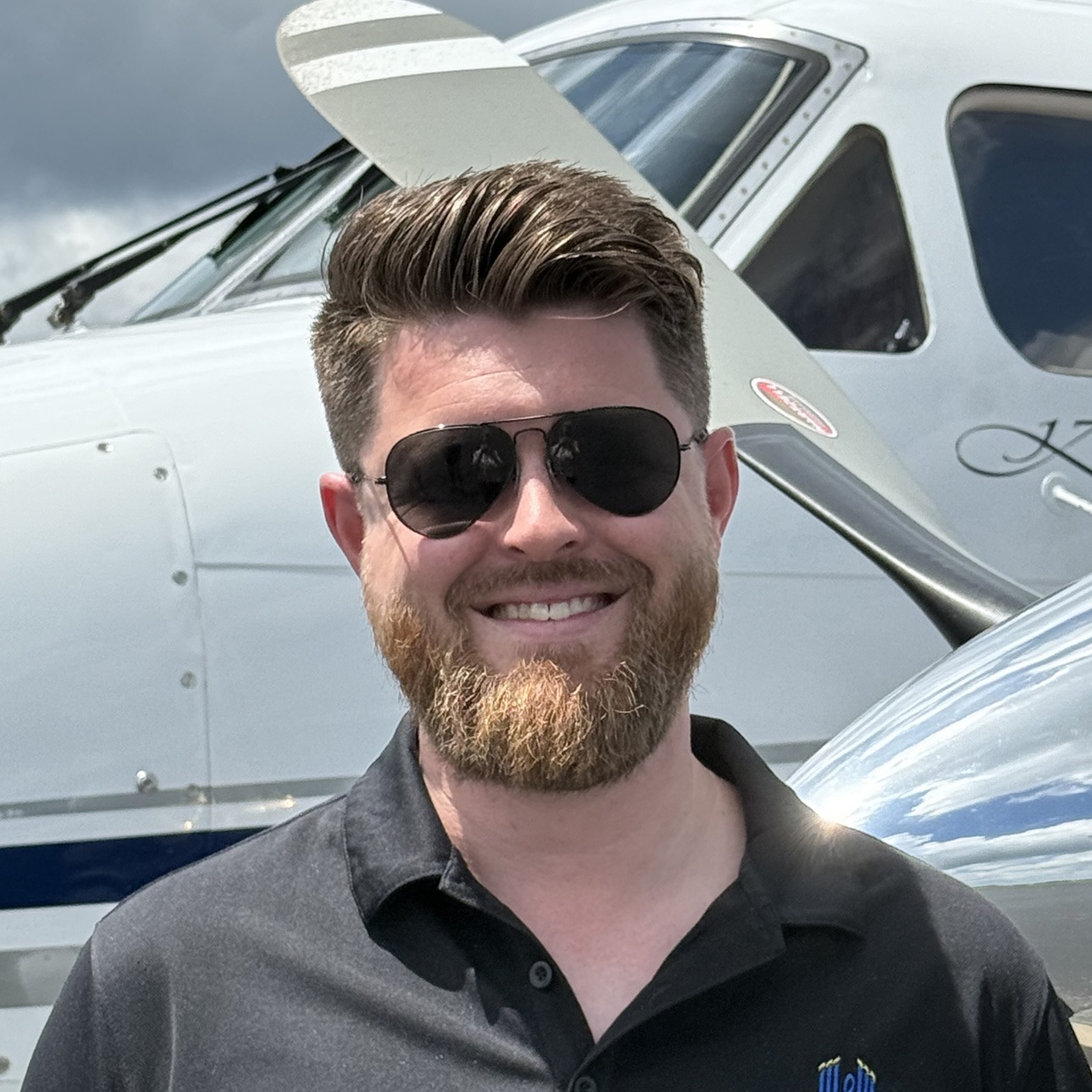 Man with a beard and sunglasses smiling in front of a small white propeller aircraft.